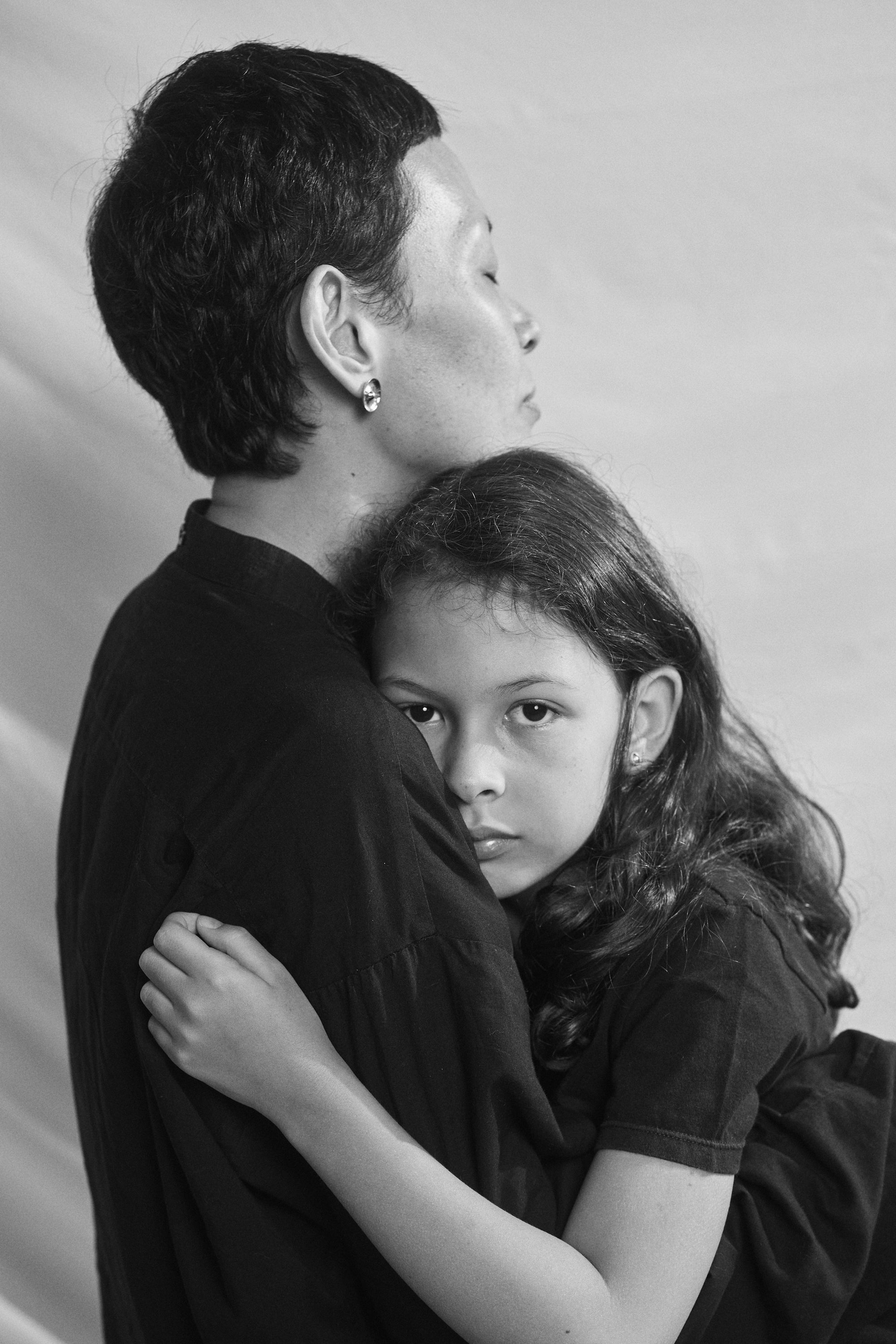 Black and white portrait of a mother lovingly embracing her daughter, both wearing Carina Hardy fine yellow gold jewelry, for mother's day. View the collection today.