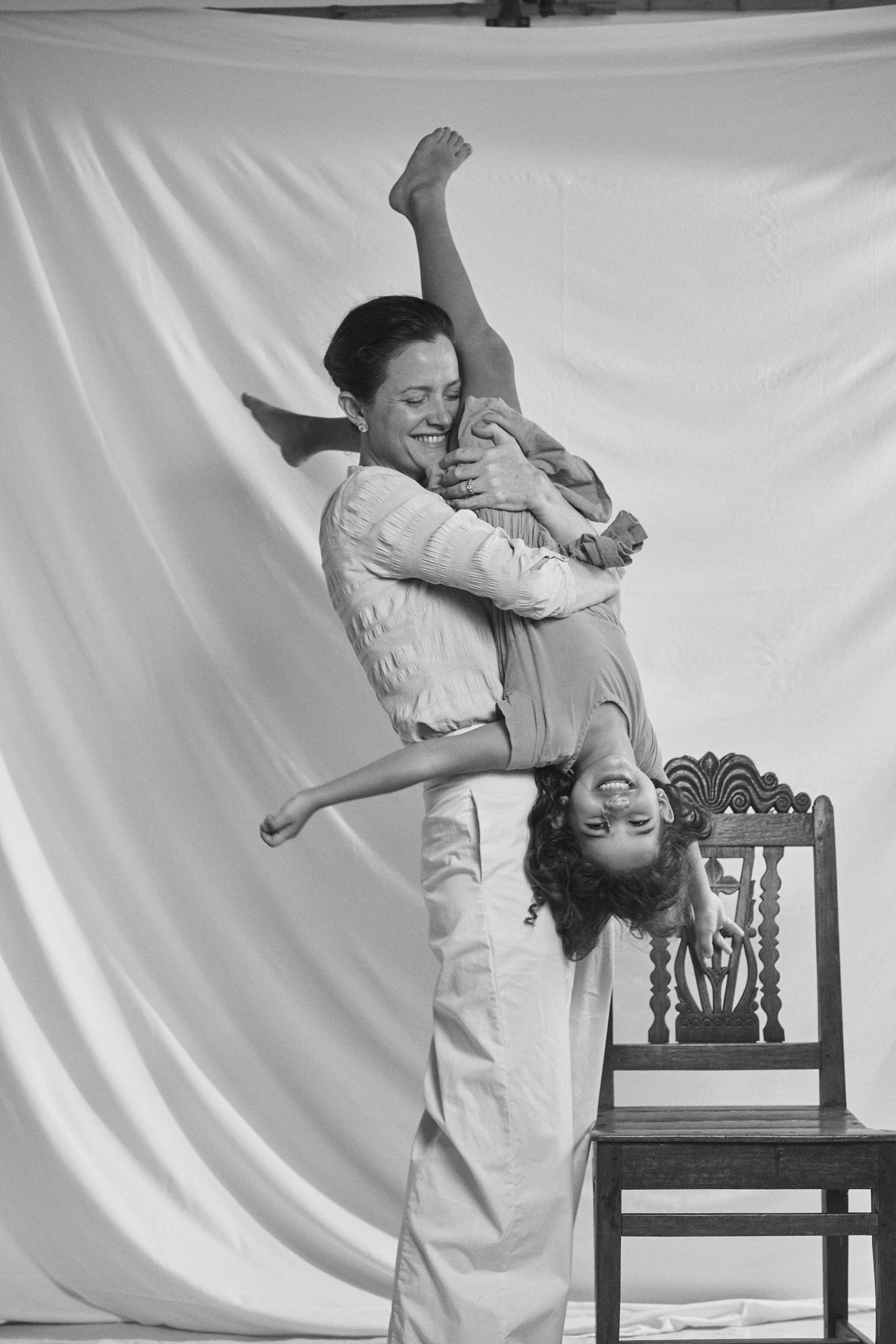 Playful black and white photo of a mother lifting her child in the air, both adorned with Carina Hardy fine yellow gold jewelry. Family moments with our pieces. View the collection today.