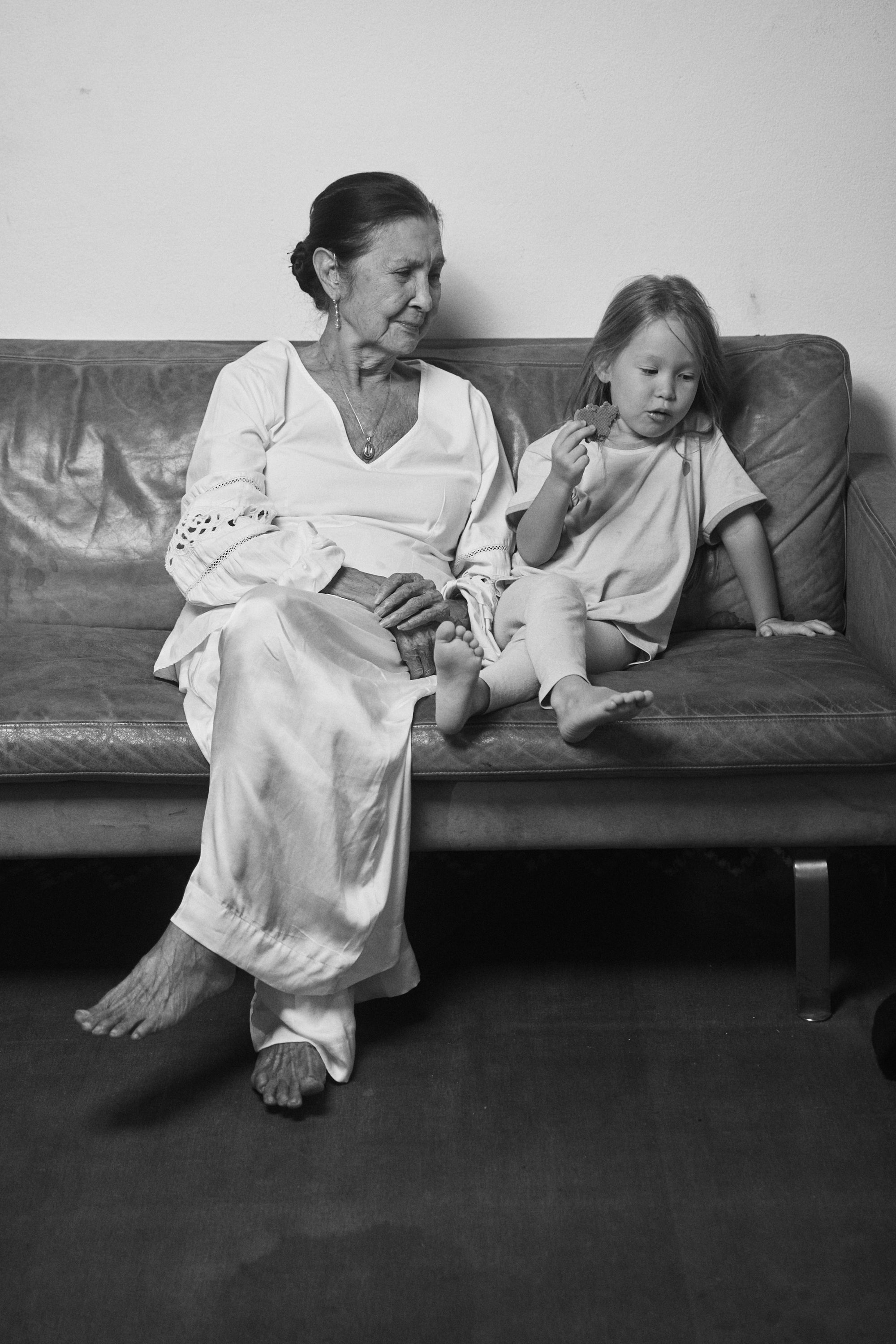 Black and white image of a grandmother and grand-daughter sitting on a couch, both wearing Carina Hardy fine yellow gold jewelry. Celebrating family moments. View the collection today.