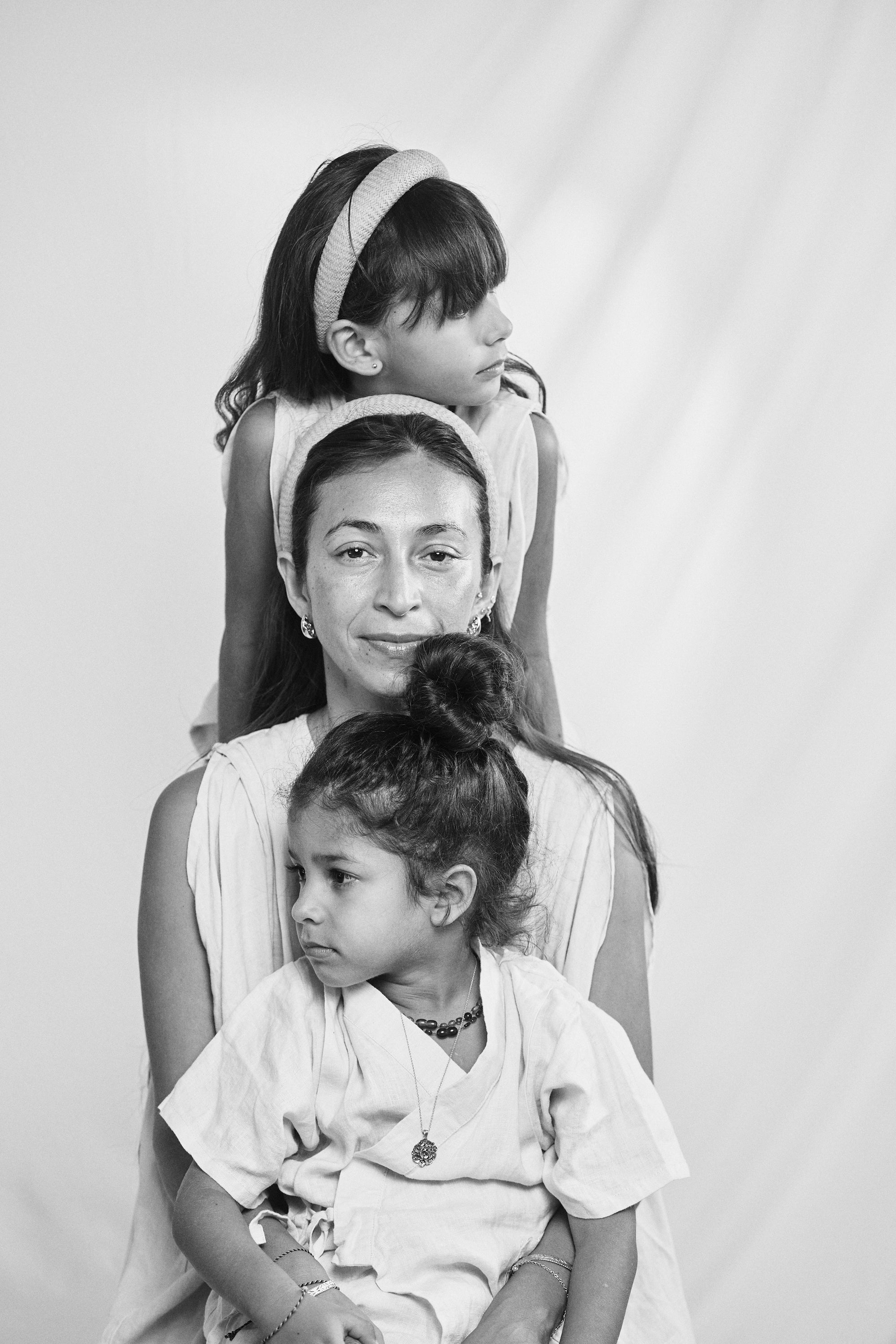 Charming black and white portrait of a mother and her two daughters sitting together, showcasing Carina Hardy yellow gold fine jewelry. View the collection today.