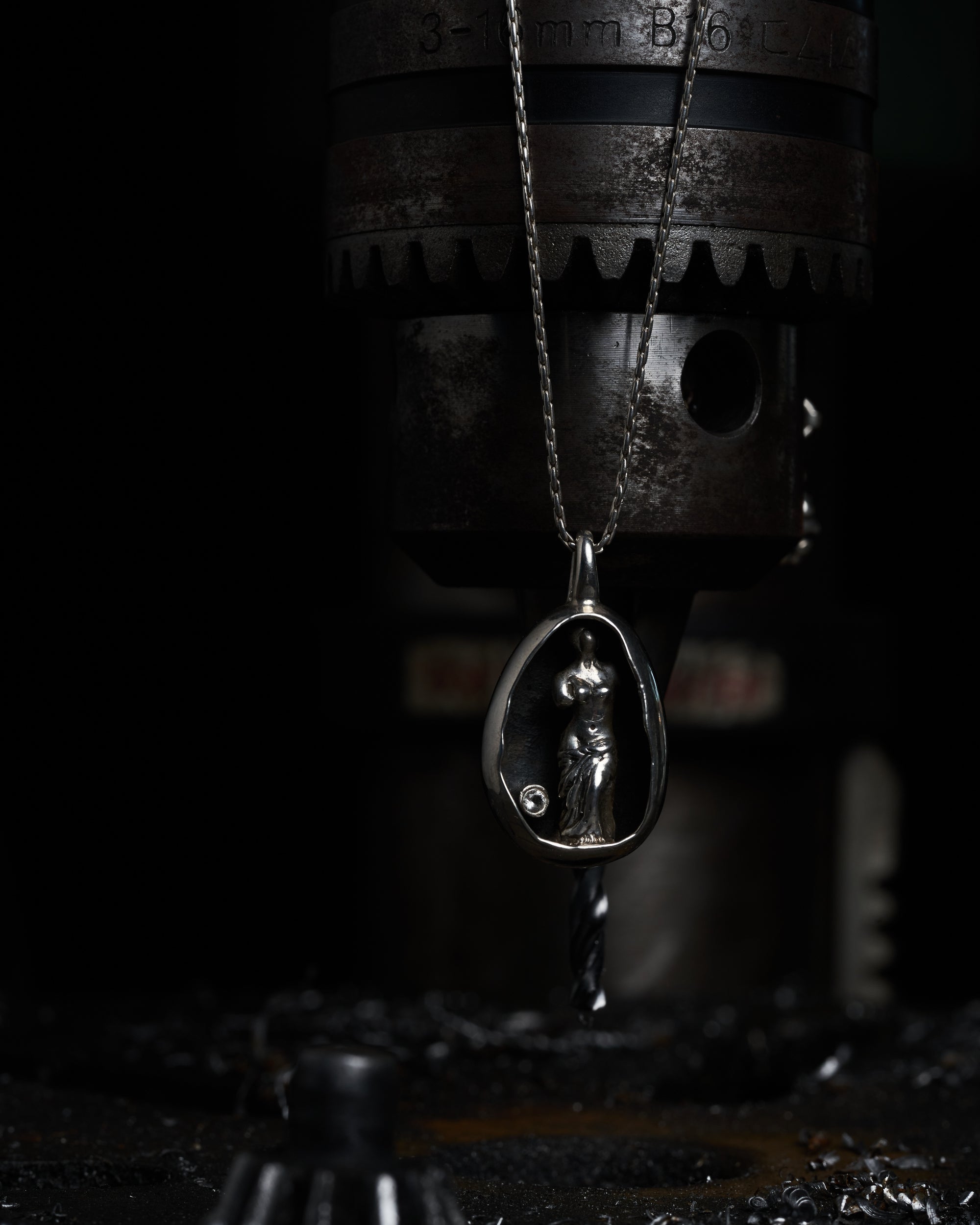 Close-up of a Carina Hardy precious sterling silver Venus de Milo pendant hanging from a machine. The contrast of darkness and light emphasizes the intricate design and meticulous craftsmanship. View the collection today.