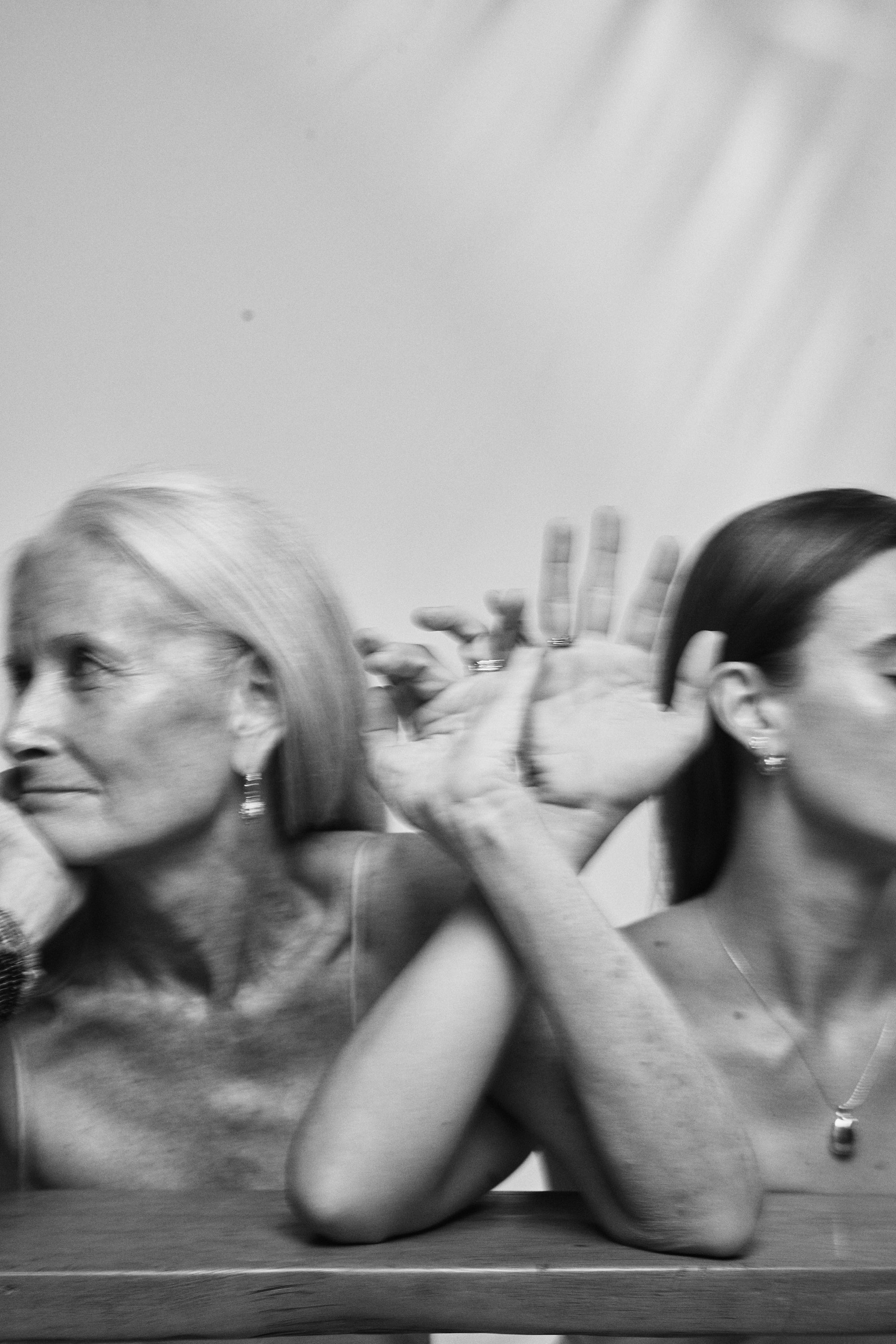 Joyful black and white image of two women entangled by the fingers together, both wearing Carina Hardy yellow gold fine jewelry. View the collection today.