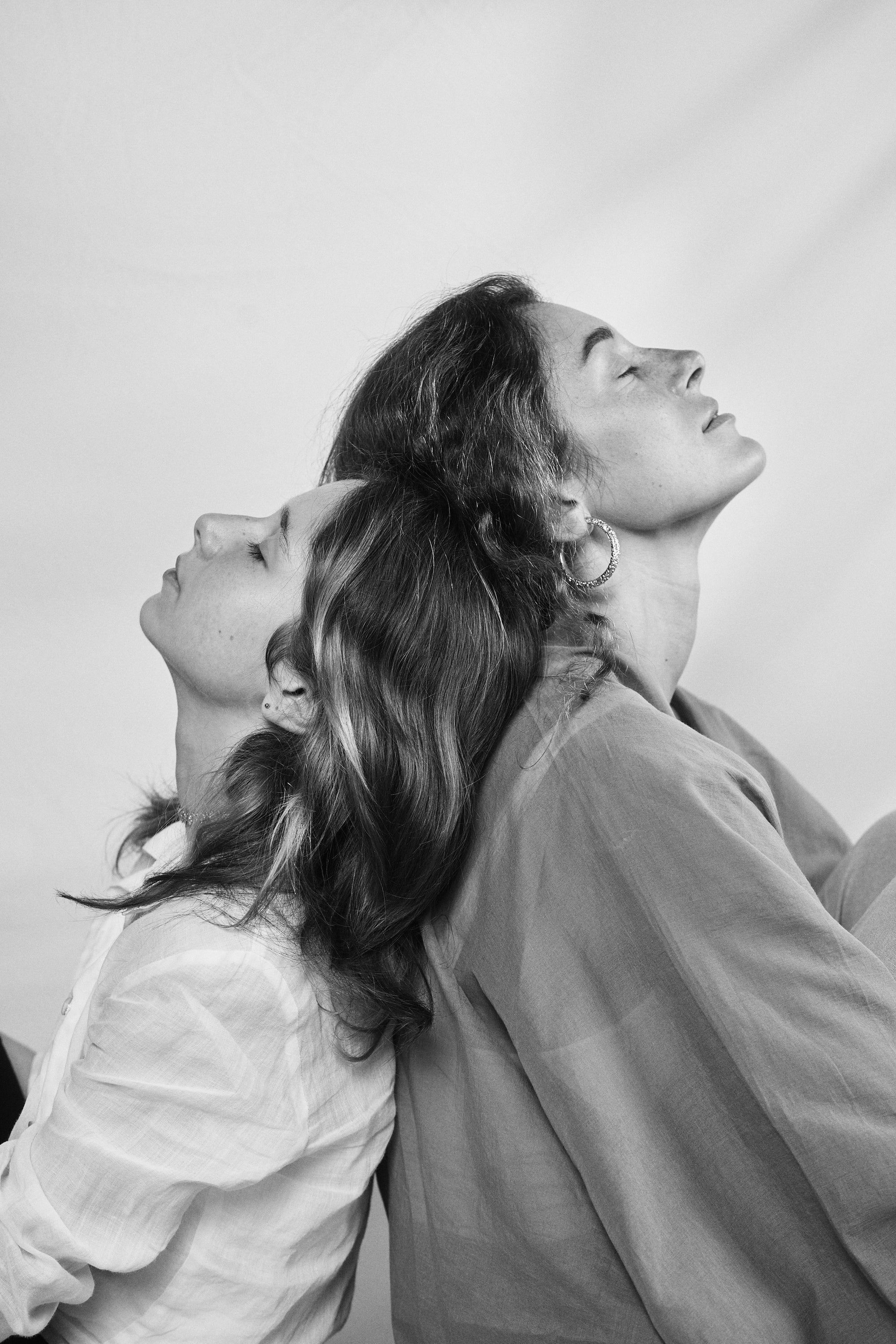 Black and white portrait of a mother and daughter leaning back-to-back, adorned with Carina Hardy fine yellow gold jewelry, their bond and style sweetly felt. View the collection today.