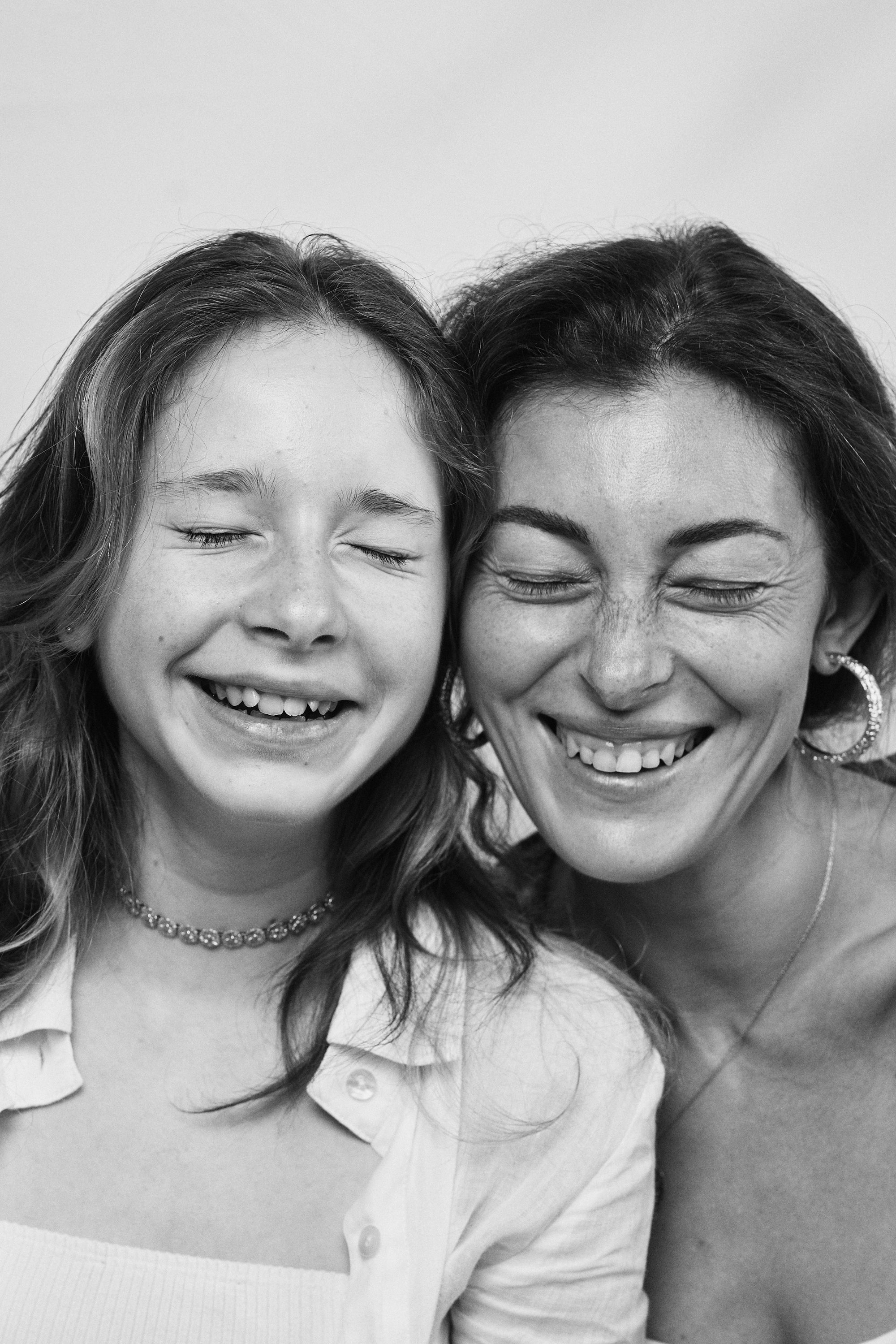 Joyful black and white portrait of mother and daughter smiling, both adorned with Carina Hardy fine yellow gold jewelry. Celebrate motherhood, view the collection.